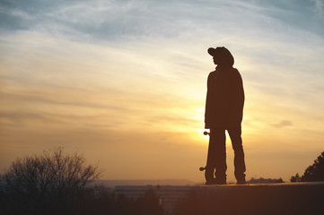 The teenager in a sweatshirt and a cap standing with a board in the city against the backdrop of the urban sunset. Silhouette photo of extreme