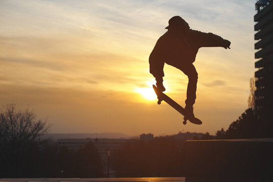 The teenager in a sweatshirt and a cap jumps with a board in the city against the backdrop of the urban sunset. Silhouette photo of extreme