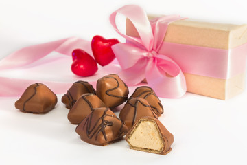 Chocolate candy on a blurry background of two red hearts and a gift in a paper box with a pink ribbon and bow on a white background. Shallow depth of field.