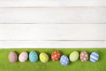Colorful handmade Easter eggs lie on a green lawn on a white wooden background. Flat lay style