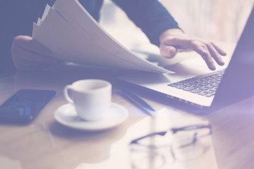 Businessman working at sunny office on laptop while sitting at the wooden table.Man holding paper documents in hands.Selective focus.Blurred background,visual effects.