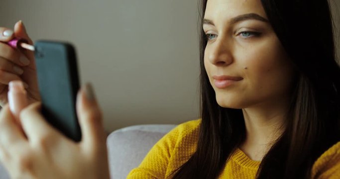 Close up beautiful woman applying lipgloss, lipstick on her lips while sitting on the couch at home. Applying make up. Slow motion