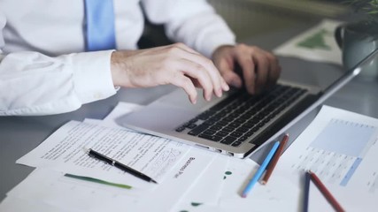Close up of a man wearing a blue tie writing down numbers and typing at his laptop keyboard. Locked down real time close up shot - Powered by Adobe