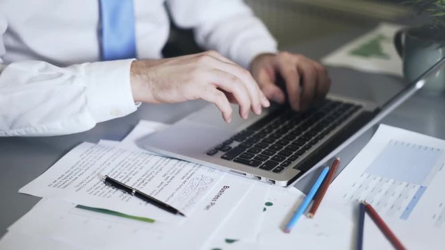 Close up of a man wearing a blue tie writing down numbers and typing at his laptop keyboard. Locked down real time close up shot