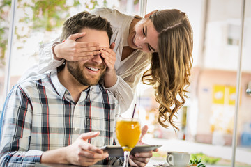 Young smiling woman closing eyes of her boyfriend in cafe