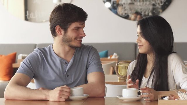 Two young friends of different genders chatting at the cafe. Handsome caucasian guy and pretty asian girl looking at each other. Attractive brunette woman touching her hair