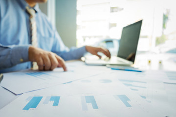 Handsome young businessman working with laptop while sitting on the desk in office and financial statistic graph, blurred background