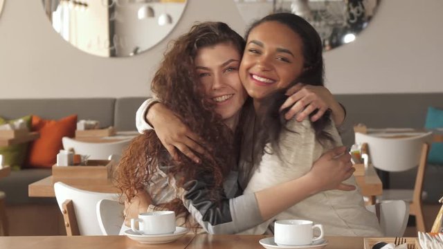 Two pretty girls showing how they like each other at the cafe. Attractive caucasian woman hugging her friend at the table. Cute african american lady posing for the camera with her curly mate
