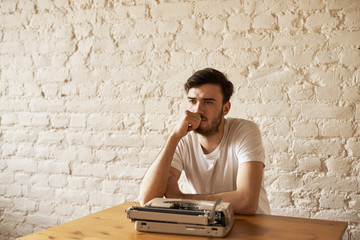Journalist sit at the table with typewriter and think about new article. Creative crisis concept. Handsome man in white t-shirt hold chin and look away with white brick wall background. Copy space - obrazy, fototapety, plakaty
