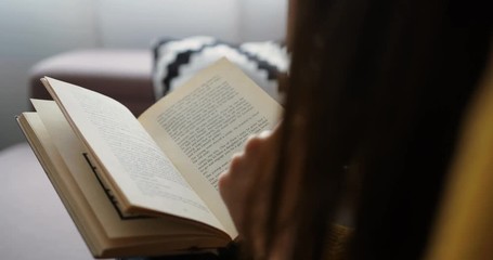 Close up woman reading interesting book while sitting on the couch at home. Close up back view. Close up book - Powered by Adobe