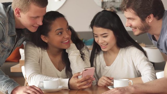 Mixed race group of friends watching something on smartphone at the cafe. Two caucasian guys standing above african american and asian girls which sitting at the table. Young people laughing about