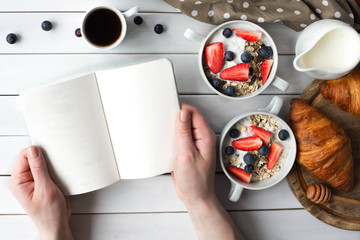 Young woman holding notebook in hands on white wooden background and have a healthy cereals breakfast with pumpkin seeds, oats,yogurt,strawberry,blueberry and coffee. Muesli bowl top view