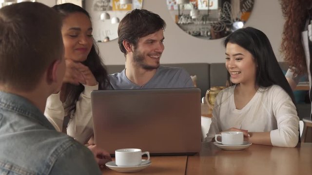 Multirace group of friends watching something on laptop at the cafe. Pretty african american girl bending over the table. Cute curly caucasian woman standing behind bearded man and brunette asian lady