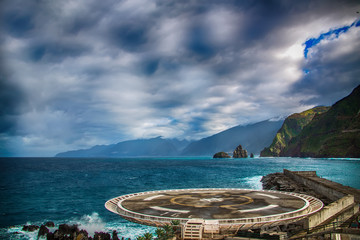 Helipad located in Porto Moniz, North of Madeira Island. In the background there are blue ocean waves, black rocks and green hills of the coastal line.