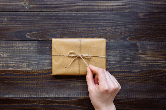 Female Hand Unpacking Gift Box Wrapped With Craft Paper And Flowers On The Wooden Table Top View. Gift For Any Holiday