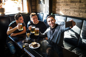 Handsome friends are drinking beer, doing selfie and smiling while resting in pub. Beer time.