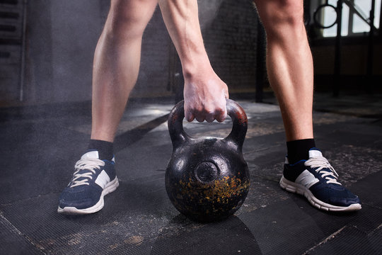 Closeup photo of young man's legs in sneackers and arm while keeping kettlebell against dark background.