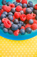 various kinds of fruit - berries raspberries, blueberries - in a blue bowl over yellow background with copy space 