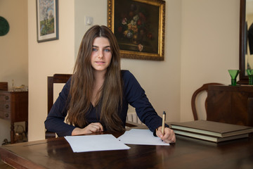 Pretty young woman with blue eyes and brown hair sitting on an elegant wooden table typing on a blank sheet in the living room of her house. She is working and dressed smartly.  