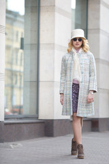 A beautiful stylish blond woman in a coat and hat and sunglasses on a city street.