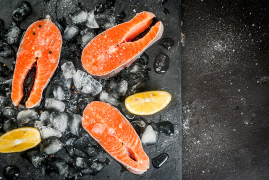 Fresh raw unprepared fish salmon trout, steaks, on a black slate board with ice, salt, pepper and lemon for cooking. Ingredients of a healthy diet. Top view copy space