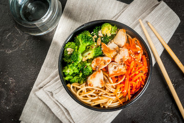 Healthy eating, diet. Buddha bowl hot salad in Asian style: noodles with soy sauce, spicy carrots, chicken, broccoli. On black concrete stone table, with chopsticks and water. Top view copy space