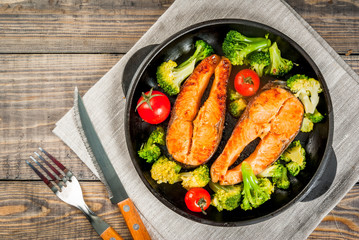 Healthy eating, diet. Baked grilled trout (salmon) with vegetable garnish - broccoli, tomatoes. In a portioned frying pan, on a wooden table. Top view copy space