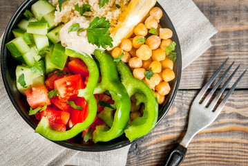 Healthy eating, diet. Vegetarian vegetable buddha bowl - couscous (quinoa), chickpeas, tomatoes, zucchini (cucumber), pepper, hummus, lemon and greens. On wooden table, with water, top view