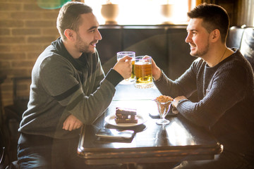 Friends drinking beer. Two cheerful young men toasting with beer and smiling while sitting together in pub