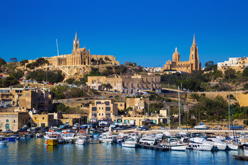 Gozo, Malta - The harbour of Mgarr with view of church Our Lady of Lourdes on top of the hill on a bright sunny summer day with blue sky
