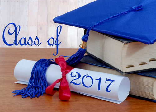 Blue graduation cap on books with rolled up diploma tied with red ribbon on wooden table. Class of 2017 text added