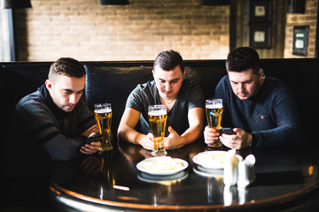 Three young men in casual clothes are talking, using a smart phone and drinking beer while sitting in pub. Phone and social network dependence