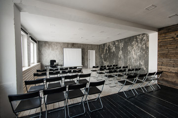 Fototapeta premium Room for lecture with a lot of dark chairs. Walls are white, loft interior. On the right there is a door. On the background there is a table with a laptop.