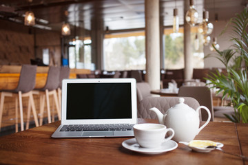 Fototapeta na wymiar Open net-book and cup of tea with lemon on table in moder cafe restaurant interior. Laptop computer with blank copy space for your text message. Freelance remote job during tea break