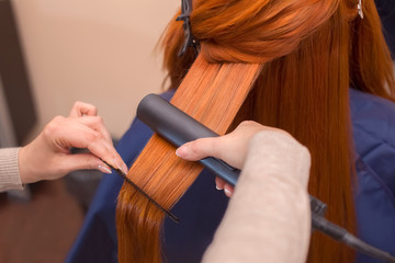 Hairdresser makes hairstyle girl with long red hair in a beauty salon. Straightening hair ironing....