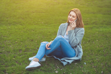 Attractive blond girl talking on mobile phone sitting on grass in nature.