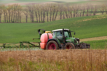 Obraz premium Farm machinery spraying insecticide to the green field, agricultural natural seasonal spring background. Tractor sprinkling pesticides against bugs on agricultural field