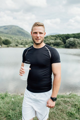 Fototapeta premium Smiling male runner standing at a pond with a bottle of water