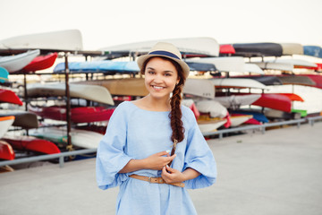 Portrait of smiling white Caucasian brunette woman with tanned skin in blue dress and straw hat, at seashore lakeshore with canoe boats on background, summer day lifestyle