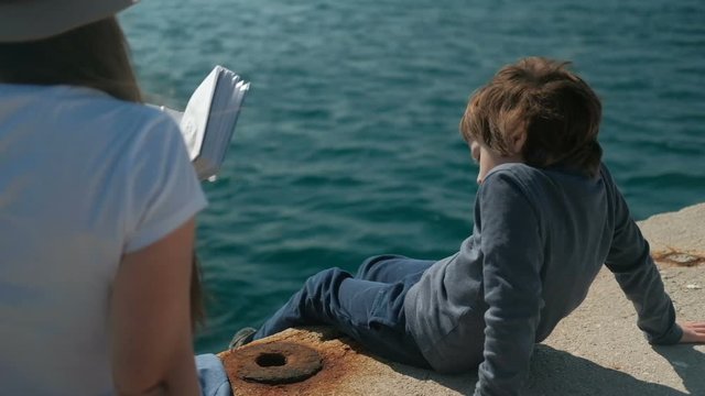 Mom reads book to son, nature scenery next to sea outdoors. Cute boy sitting on concrete pier in blue sweater, pants, black boots, swinging with feet, listening to fairy tale. Mom sits back with long