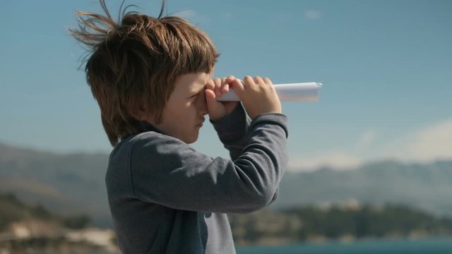 boy folded piece of paper looking into the distance like trumpet to mountains. Slow motion picture of child standing on lake holding hands with paper in cage, like telescope, closed right eye, looks