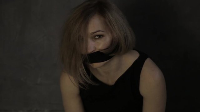 frightened crying girl gagged, sitting on a floor with tied hands. linen bag on the head of a hostage. kidnapping and violence