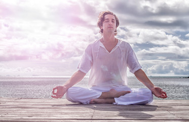 Fototapeta na wymiar Young man meditating with closed eyes on the beach, clouds in the background