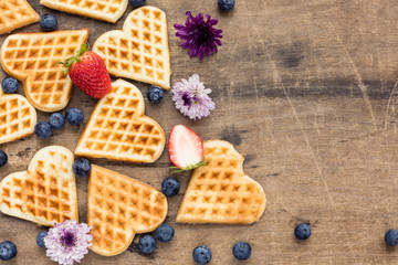 Fresh homemade heart shape waffles with blueberries and strawberry on wooden background, top view
