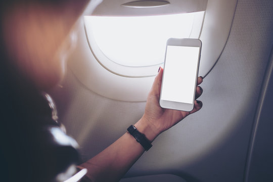 Mockup image of a woman holding and looking at white smart phone with white screen next to an airplane window with clouds and sky background