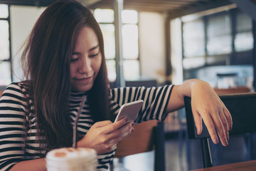 A beautiful Asian girl with smiley face holding , using and looking at smart phone in modern cafe