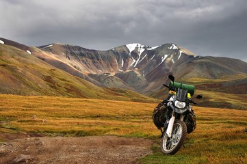 Enduro motorcycle traveler with suitcases standing on dry yellow grass meadow on the background of colorful high snow mountain and stormy dark sky with  clouds Plateau Ukok Altai Siberia Russia