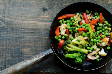 Frying pan with fried egg and a vegetable mixture: peas, broccoli, paprika and frozen broccoli on the side on natural wooden background, top view