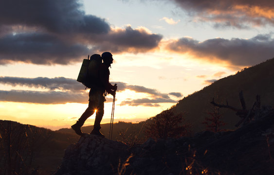 Young mountaineer standing with backpack on top of a mountain silhouette in mountains with sunset background