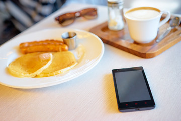 The concept of morning breakfast, mobile phone, mug of coffee and tasty pancakes on wooden desk.